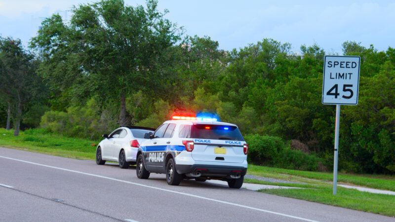 A police SUV with flashing lights has pulled over a sports car on the side of the road just past a sign denoting a 45 MPH speed limit.