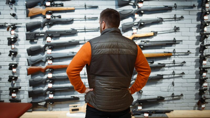 A man in an orange shirt and a vest stands with his back to the camera in front of a wall of long guns, for sale and on display.