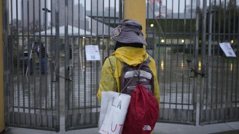 a person with a bucket hat, yellow jacket, and red backpack with a rally sign stands in front of barred doors