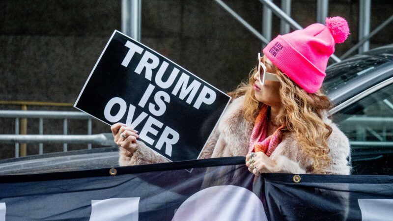 a woman outside Bragg's office yesterday wears a pink cap and holds a sign saying "Trump is over"