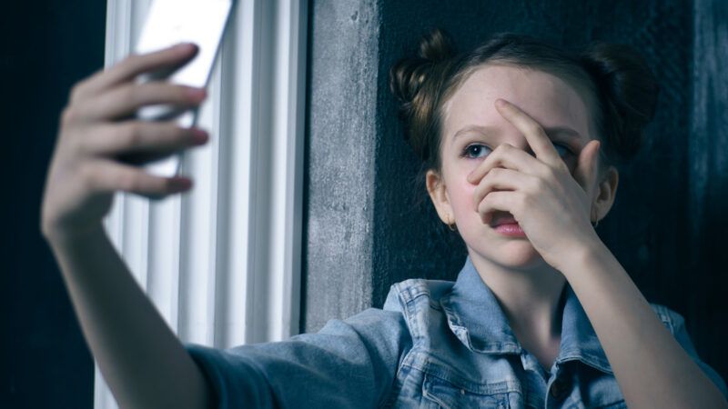 A scared young girl peeks through her fingers at her cell phone screen.