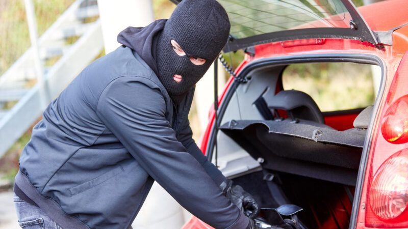A thief in a ski mask looks around before stealing something out of the back of a car's open hatchback.