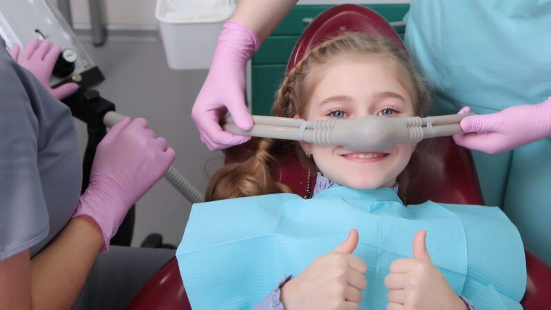 A little girl smiles and gives a thumbs-up as dental professionals set her up on nitrous oxide anesthesia.