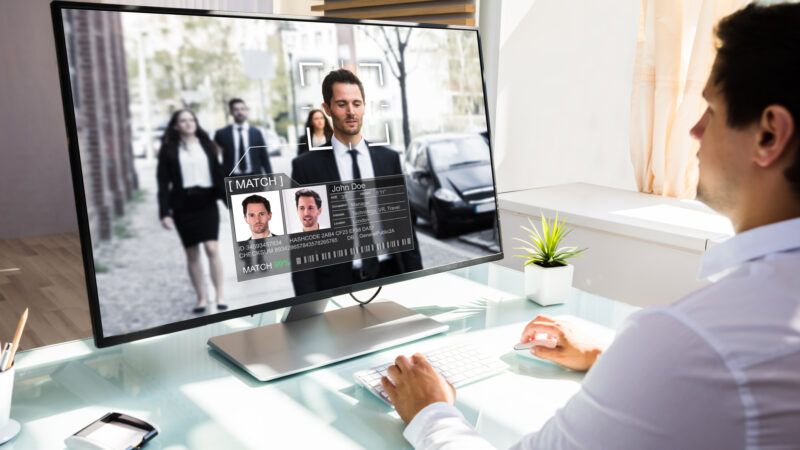 A man in a white shirt checks CCTV security footage on his computer while a facial recognition program runs.