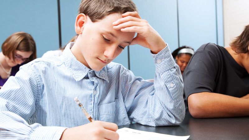 A young boy in a classroom takes a test. His hand rests on his brow as if he's thinking deeply.