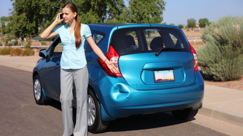 A woman stands by her dented car on the phone.