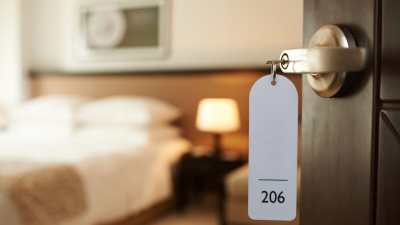 The entrance to a hotel room, with a keytag reading "206" hanging on the doorknob in the foreground and the room itself blurry in the background.