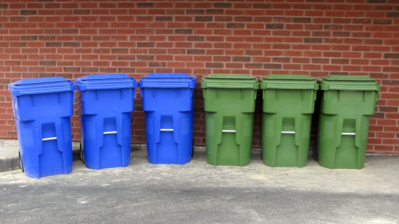 Six recycling bins, three blue and three green, lined up in a row against a brick wall