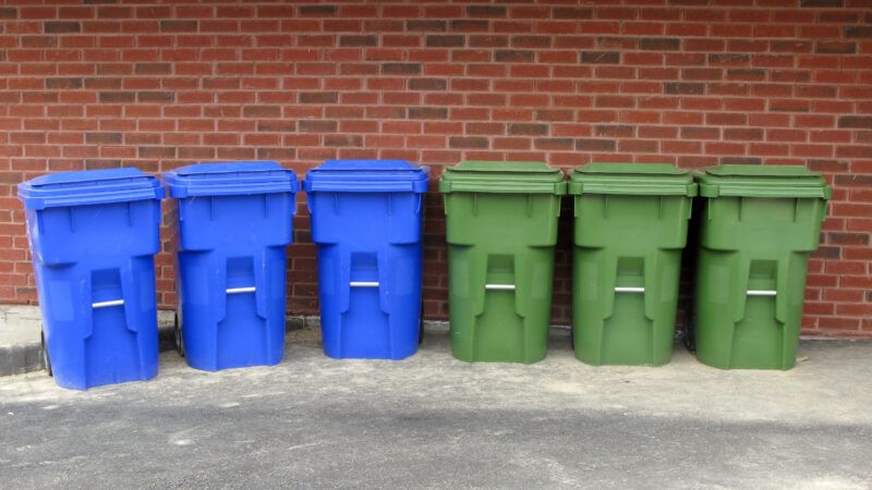 Six recycling bins, three blue and three green, lined up in a row against a brick wall | Rosamund Parkinson | Dreamstime.com