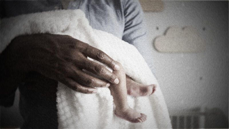 A person cradles a partially seen baby wrapped in a blanket with a nursery wall in the background