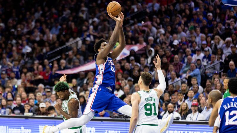 NBA player Joel Embiid takes a shot, mid-air, with players on the Boston Celtics around him.