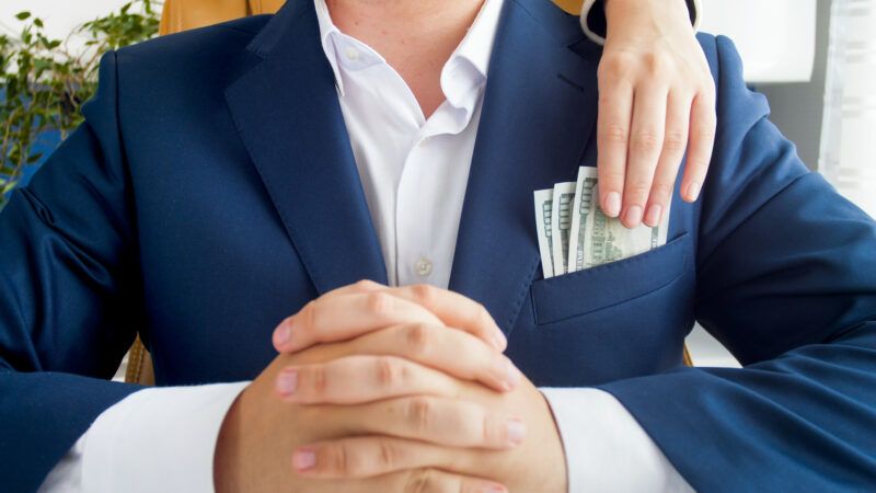 A businessman with a suit sits with hands folded in front of him, as a hand reaches over his shoulder and steals cash out of his pocket.