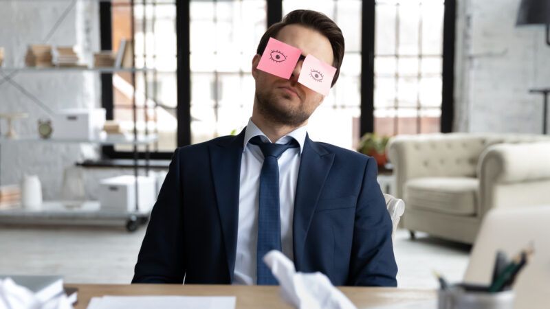 A man in a business suit sits at his desk, asleep but with Post-It notes on his eyes with eyes drawn on. | Fizkes | Dreamstime.com