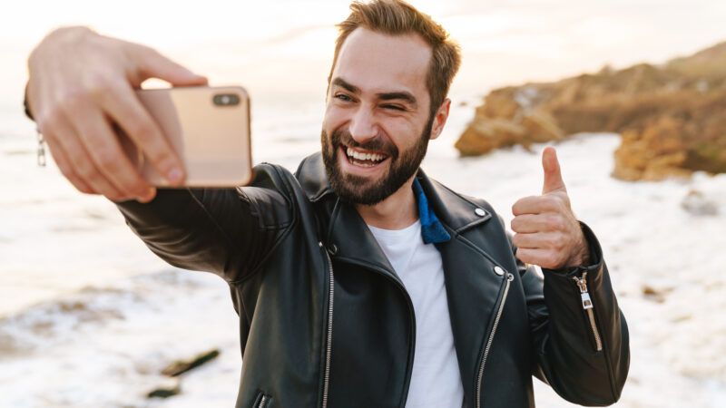 A man in a leather jacket walking along the beach takes a selfie on his phone.