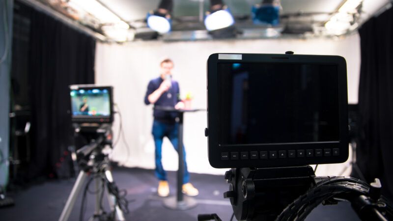 Journalist in a makeshift studio with monitors in the foreground.