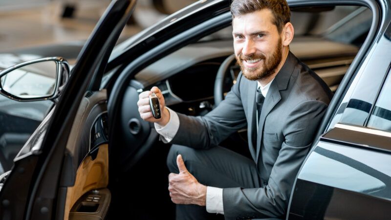 A businessman sits in the front seat of a luxury car in the showroom, holding the key and giving a thumbs-up. | Rosshelen | Dreamstime.com