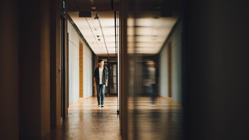 Teen walking down school hallway next to blurry mirror image of teen walking down school hallway | Photo by <a href=https://reason.com/2023/08/11/suicide-down-8-4-among-teens-and-young-adults/