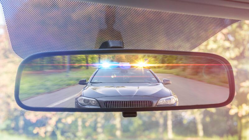 Police with flashing lights seen through a rearview mirror. | Vchalup | Dreamstime.com