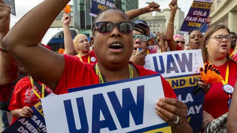 A woman wearing sunglasses holds up a UAW protest sign with other protesters who are also wearing red shirts. | Jim West/ZUMAPRESS/Newscom