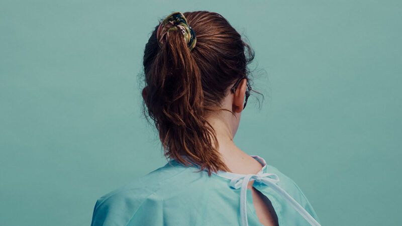 A woman in a blue gown with a ponytail faces towards a blue wall | <em>The Retrievals</em>/Serial Productions and The New York Times
