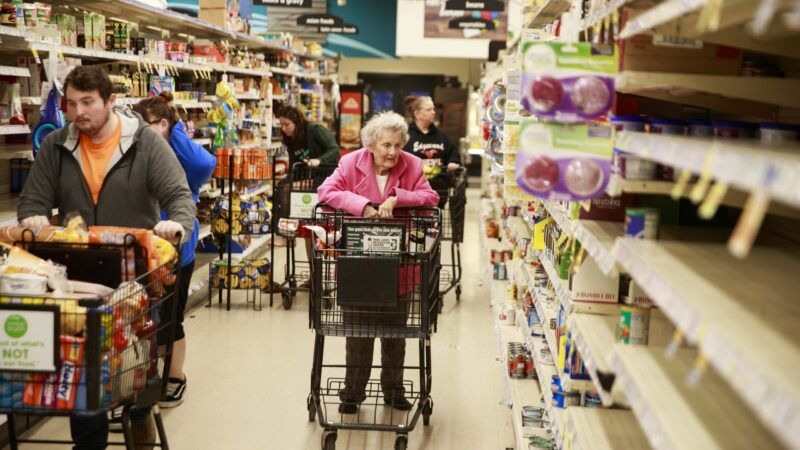 A man in a hoodie walks down a grocery store aisle with a cart of products, while an older woman wearing pink pushes an empty cart. The aisle has some products but also some bare shelves. | Jeremy Hogan/Polaris/Newscom