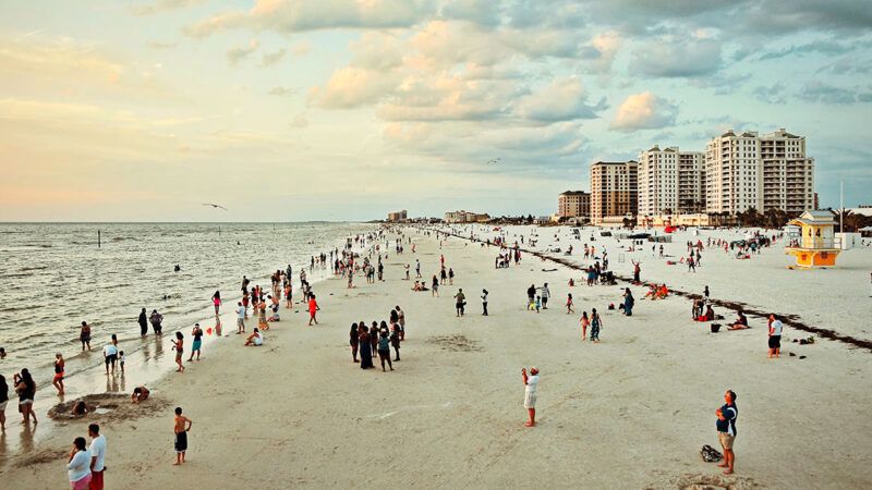 Beachgoers swim and hang out along a broad coastline with buildings in the background | Photo: robert-linder/Unsplash