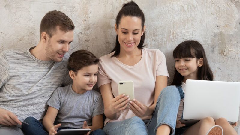 A family gathers around a cell phone and a laptop | Photo 156764429 © Fizkes | Dreamstime.com