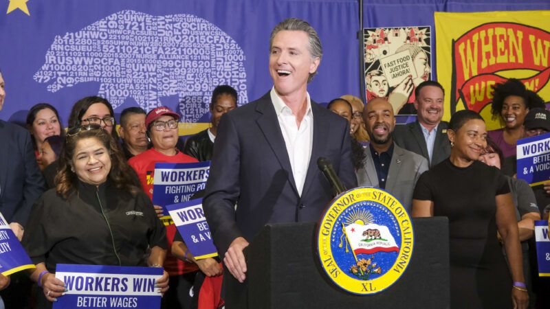 Gavin Newsom speaking at a lectern with people behind him holding signs that say "workers win" | Ringo Chiu / SOPA Images/Sipa USA/Newscom
