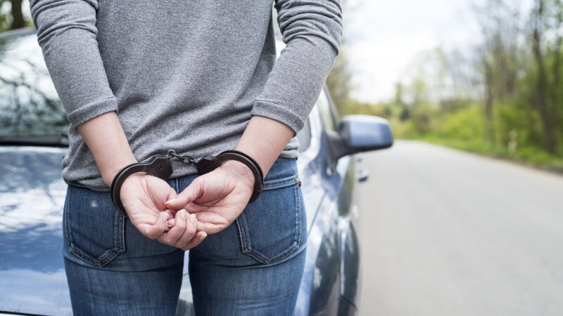 A woman stands in front of a police car on the side of the road, handcuffed behind her back. | Piotr290 | Dreamstime.com
