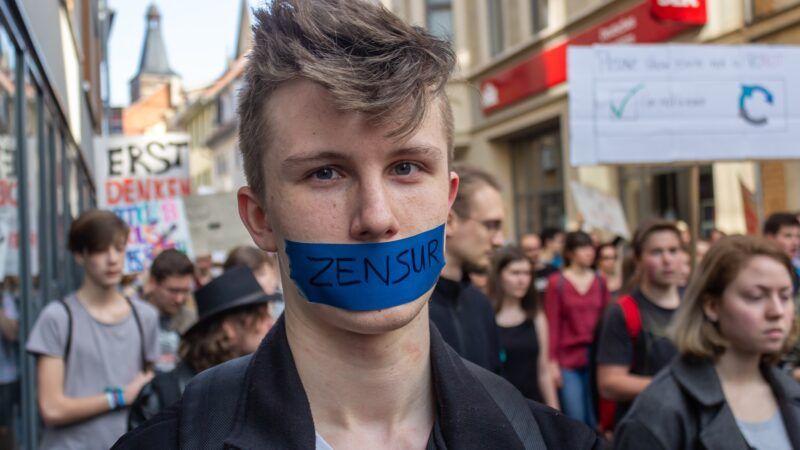 A young German man in a crowd, with blue tape over his mouth that says 'ZENSUR' (censorship). | Wirestock | Dreamstime.com