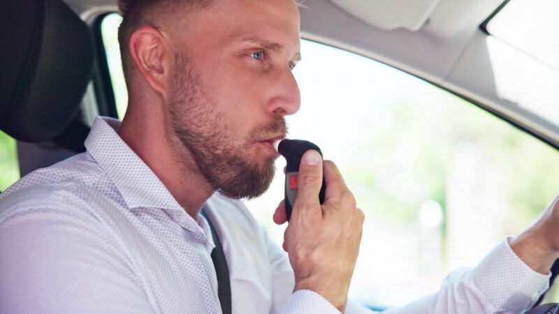 A man in the drivers seat of a car blows into a breathalyzer. | Andrey Popov | Dreamstime.com