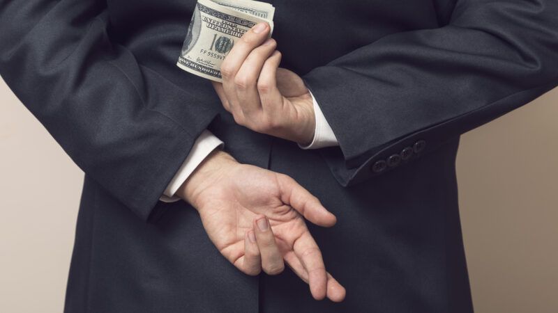A man in a business suit holds a wad of cash behind his back while crossing his fingers on his other hand. | Vladans | Dreamstime.com