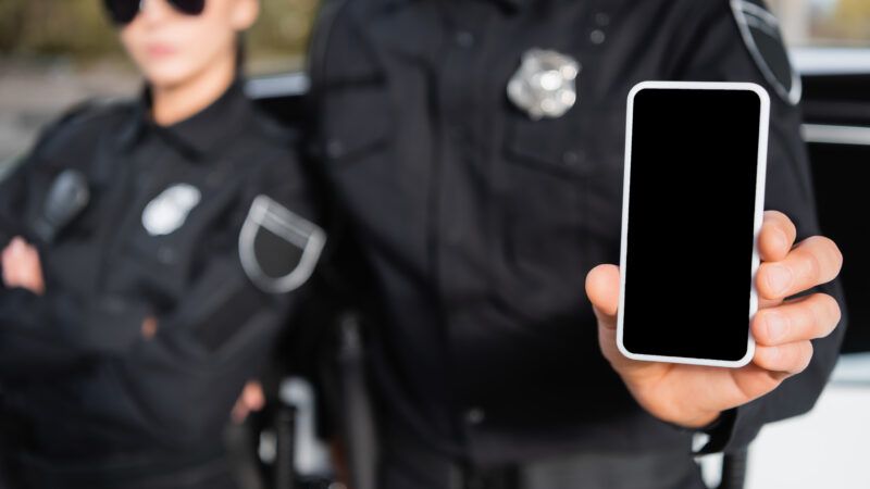 Two police officers stand in a blurred background, while one holds up a smartphone with a blank screen | Lightfieldstudiosprod | Dreamstime.com
