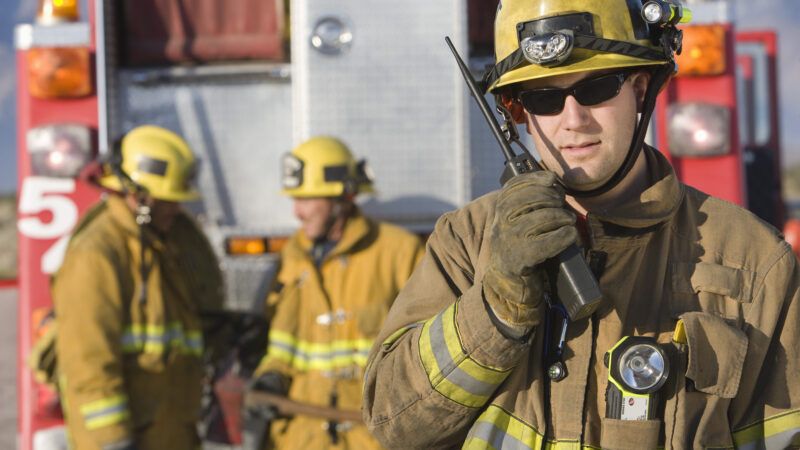 A firefighter talks into a handheld radio as two others lean against a fire engine in the background. | Photographerlondon | Dreamstime.com