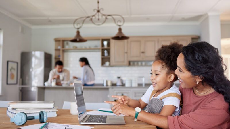 A mother and daughter crowd around a laptop at the kitchen table, as part of a homeschool setup. | Yuri Arcurs | Dreamstime.com