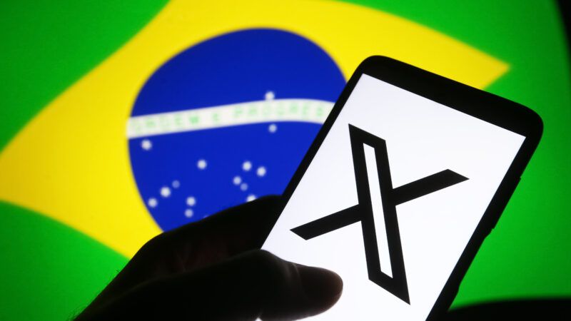 A hand in silhouette holds a smartphone displaying the logo of X (formerly Twitter), against the backdrop of the flag of Brazil. | Pavlo Gonchar/ZUMAPRESS/Newscom