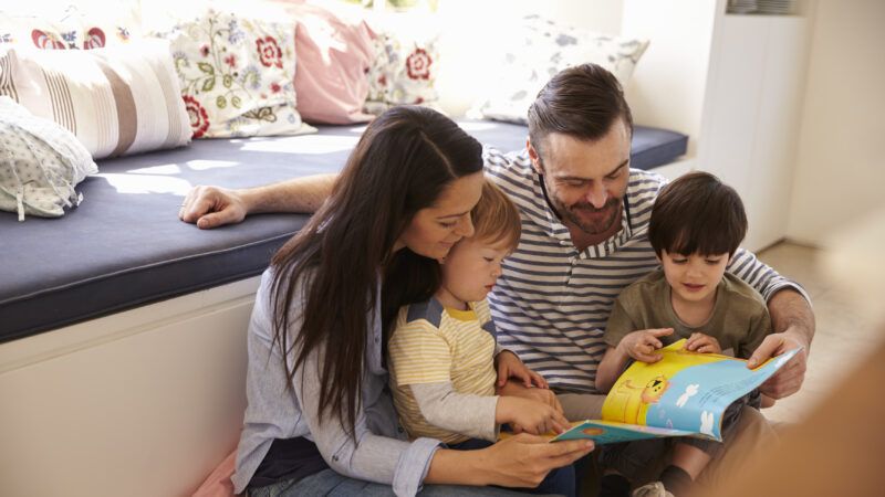 A family with father, mother, and two children sit on the floor reading a book together. | Monkey Business Images | Dreamstime.com