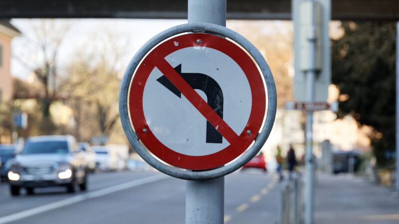 An old damaged traffic sign on the side of a busy street, signifying "No Left Turn," with a Left-Turn arrow and a red circle and line through it. | MediaCastle | Dreamstime.com