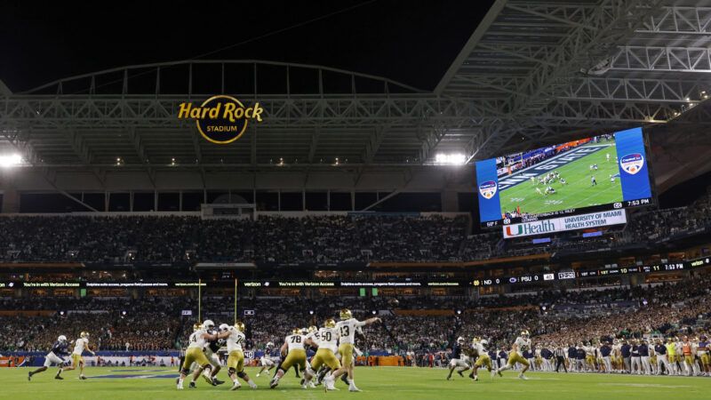 An overview of Hard Rock Stadium during a football game, with many Notre Dame football players on the ground in white jerseys and gold helmets, and a large videoboard in the right side of the stadium. | Joe Robbins/Icon Sportswire 573/Joe Robbins/Icon Sportswire/Newscom