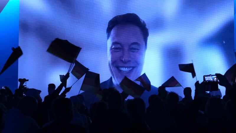 Elon Musk on a large screen in front of an audience in silhouette | 	Sebastian Willnow/dpa/picture-alliance/Newscom