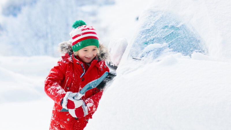 A child in a heavy red coat and hat scrapes snow and ice off a car. | Famveldman | Dreamstime.com