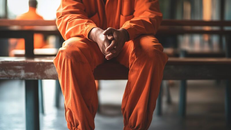 A prison inmate in an orange jumpsuit sits on a bench with hands clasped. | Frank Gaertner | Dreamstime.com