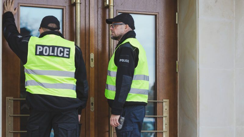 Two police officers in neon "POLICE" vests stand at a building's entrance, in front of closed doors. | Katarzyna Bialasiewicz | Dreamstime.com