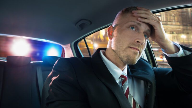 A man in a suit, in the driver's seat of his car, puts his hand to his head as he sees police lights through the back window. | Andrey Popov | Dreamstime.com