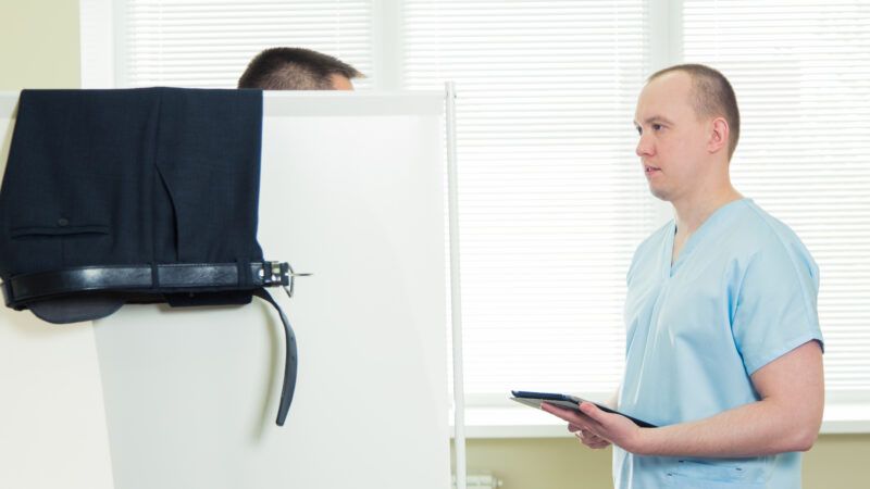 A doctor in blue scrubs speaks to a patient, who is concealed behind a divider, in a doctor's office. | Aleksey Satyrenko | Dreamstime.com