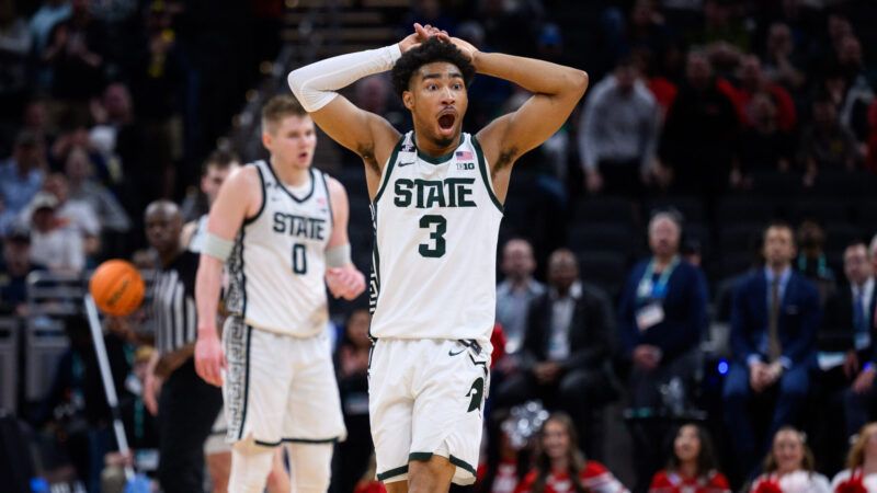 A basketball player in white Michigan State jersey holds his hands over his head with his mouth agape. There is another Michigan State player in the background, as well as a bouncing basketball and Wisconsin cheerleaders. | Zach Bolinger/Icon Sportswire DAL/Zach Bolinger/Icon Sportswire/Newscom