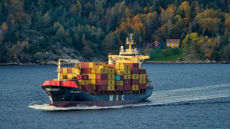 A cargo ship loaded with containers sails down a body of water with trees seen in the background on shore | Photo by Vidar Nordli-Mathisen on Unsplash