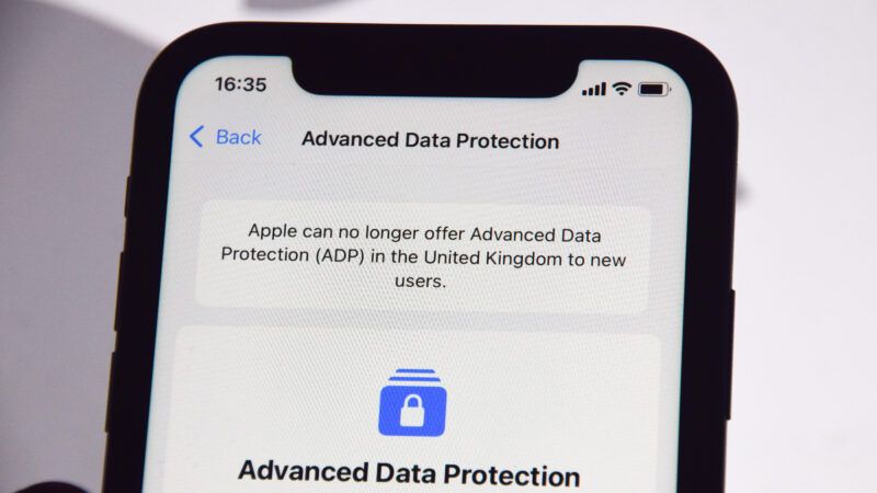 An iPhone is held against the backdrop of a giant Apple logo. The phone's screen is open to the Advanced Data Protection page. A notice at the top says, "Apple can no longer offer Advanced Data Protection (ADP) in the United Kingdom to new users." | Vuk Valcic/ZUMAPRESS/Newscom