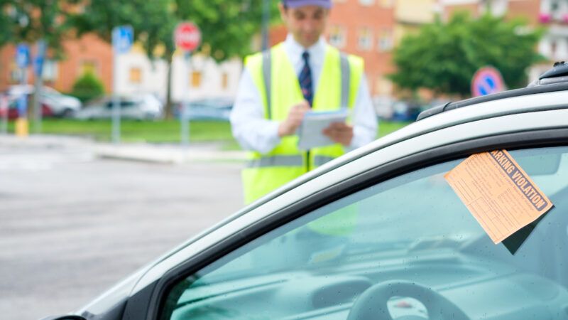 A car has a "PARKING VIOLATION" citation wedged into the driver's window. In the background, a parking attendant in a neon vest writes up the information. | F01photo | Dreamstime.com