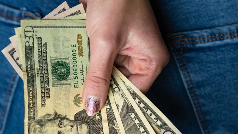 Close-up of a woman's hand behind her back, clutching fanned-out cash in 20s and 50s. | Vlad Ispas | Dreamstime.com